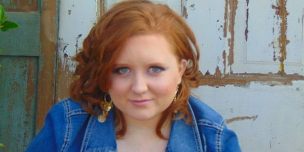 Red-haired woman in denim jacket posing against a rustic wooden backdrop.