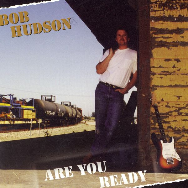 Man leaning against a wall with a guitar, train in background, album cover for Bob Hudson's 'Are You Ready'.