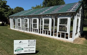 A partially constructed greenhouse with a green metal roof and white frames on a grassy lawn.