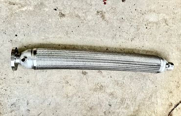 A metallic braided flexible pipe with flanged and threaded ends on a concrete surface.