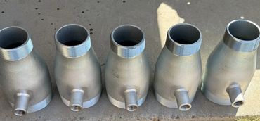 Five metallic pipe fittings with threaded ends and side outlets arranged on a concrete surface.