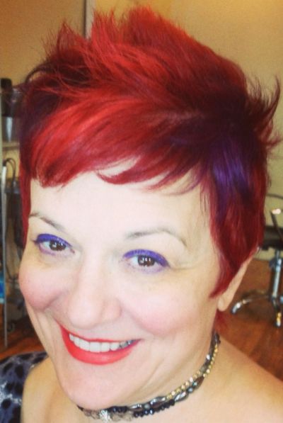 Woman with bright red hair and purple eye makeup smiling.