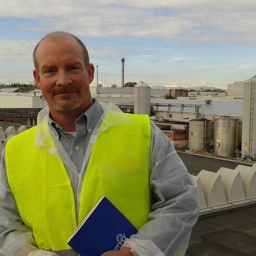 Man in a yellow safety vest standing on an industrial rooftop holding a blue notebook.