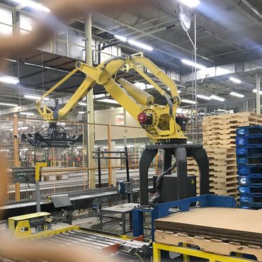 Yellow industrial robotic arm working in a factory with stacked pallets.