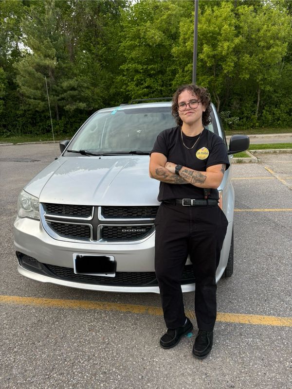 Person with tattoos stands confidently in front of a silver Dodge car in a parking lot.
