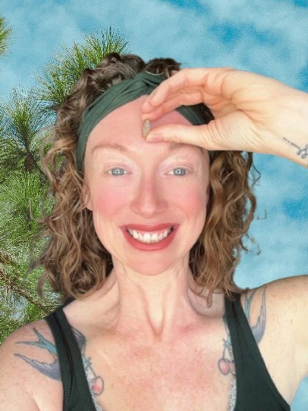 Smiling woman with curly hair holding a small pine cone against a blue sky background.