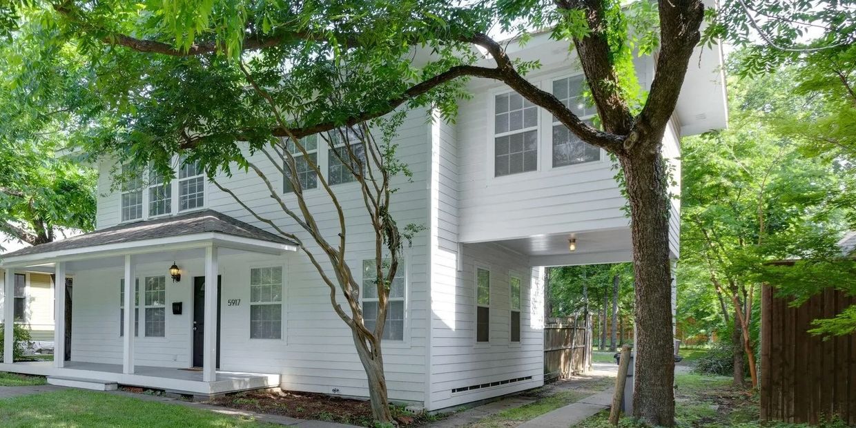 White two-story house with a covered porch and lush green trees.