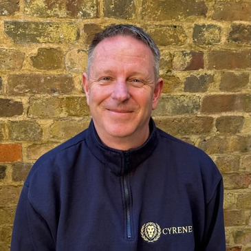 Smiling man in a navy blue Cyrene jacket against a brick wall.