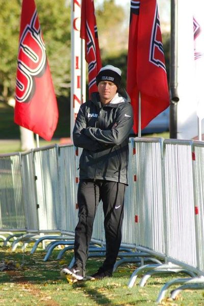 Man in sportswear standing with arms crossed near red flags and metal barriers outdoors.