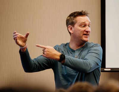 A man in a blue shirt gestures expressively while speaking indoors.