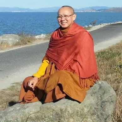 A monk in orange robes sits by a seaside path under a clear blue sky.