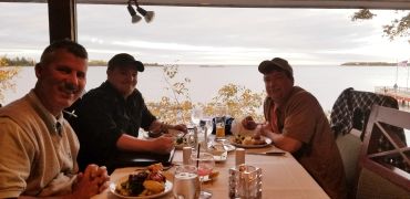 Three men enjoying a meal together by a lake or large body of water.