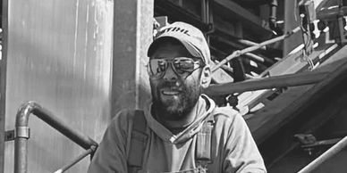 Smiling man in workwear and cap at an industrial site.