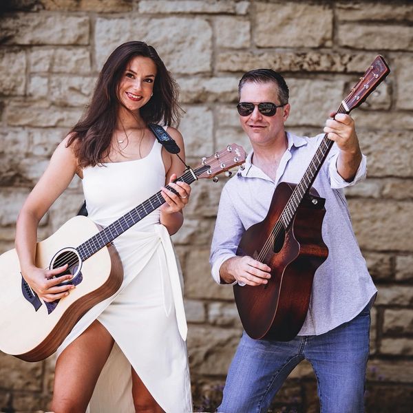 A smiling woman and man playing guitars against a stone wall.