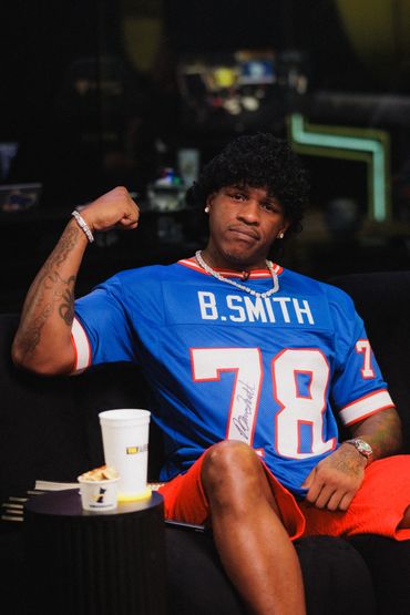 Man in blue B. Smith jersey flexes arm while sitting on black couch.