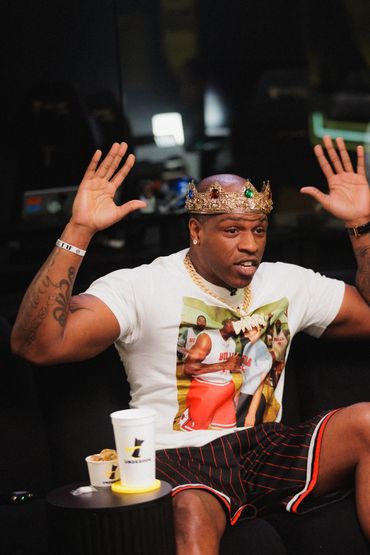 Man wearing a crown and graphic t-shirt with arms raised, sitting on a couch.