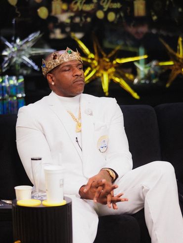 Man in a white suit and jeweled crown sitting thoughtfully on a black couch.