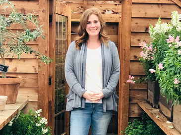 Jill Baldwin standing in front of a greenhouse with blooming plants on shelves on either side of her