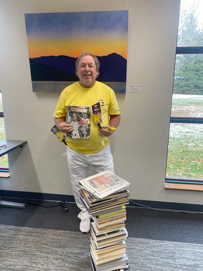 Man in yellow shirt holding magazines, standing beside a tall stack of magazines.