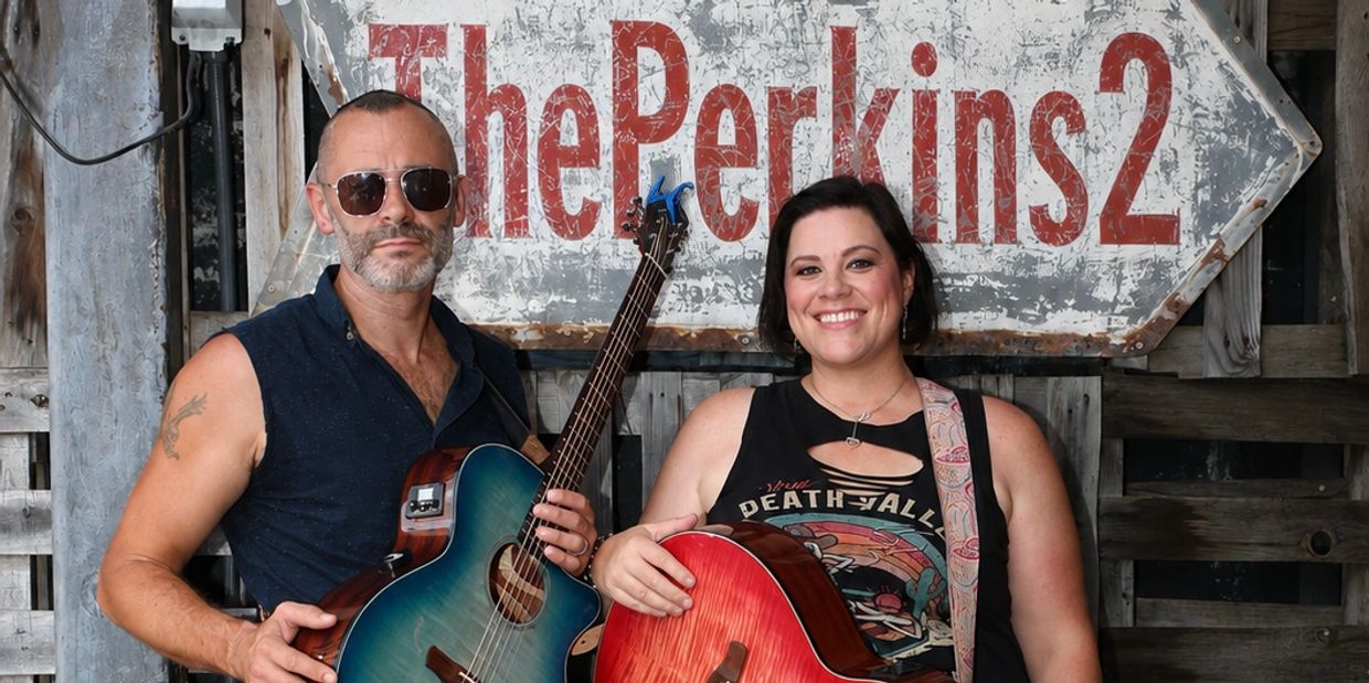 Two musicians holding guitars in front of a The Perkins 2 sign.