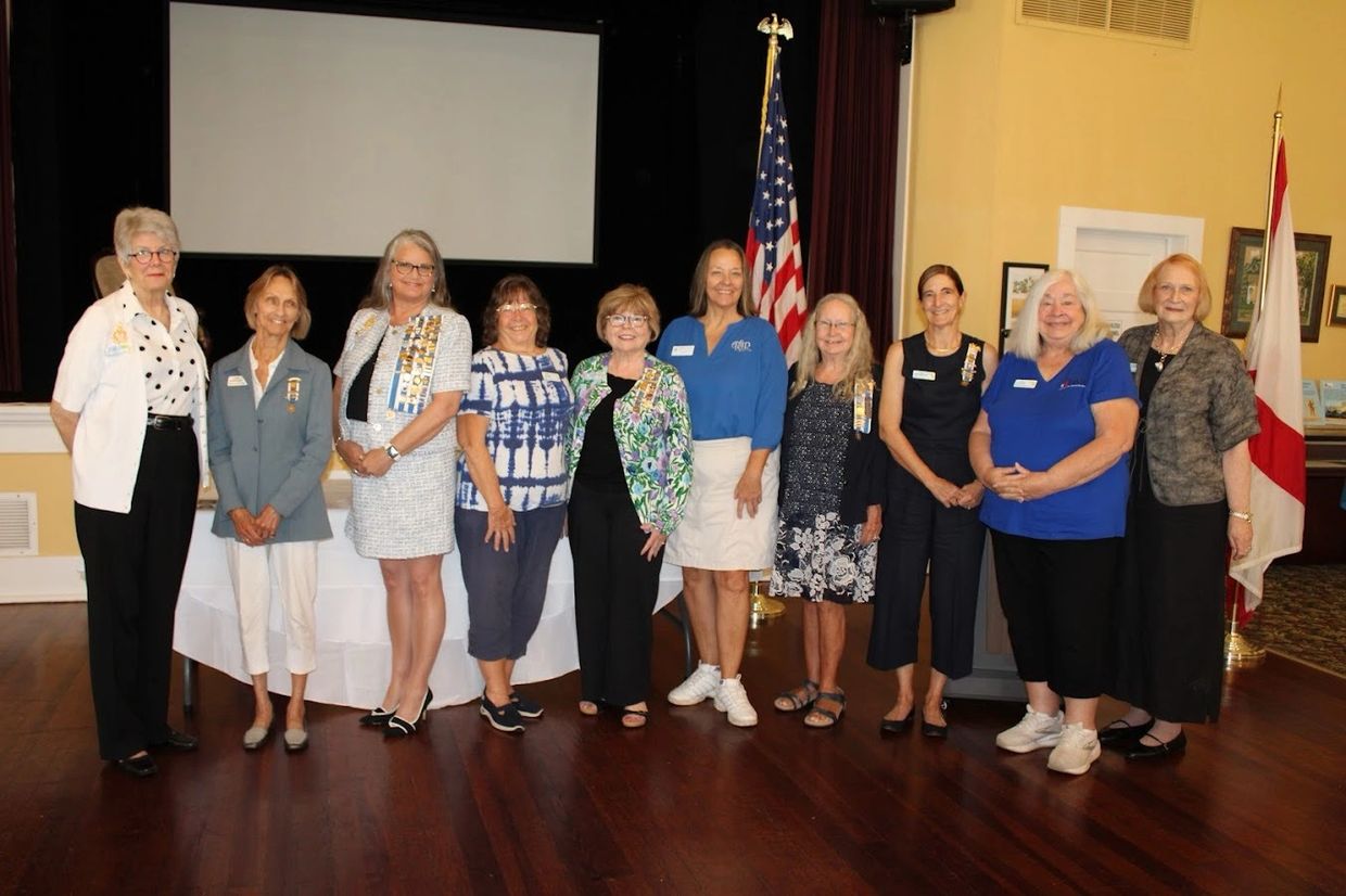 Group of ten women standing indoors with American and other flags in the background.
