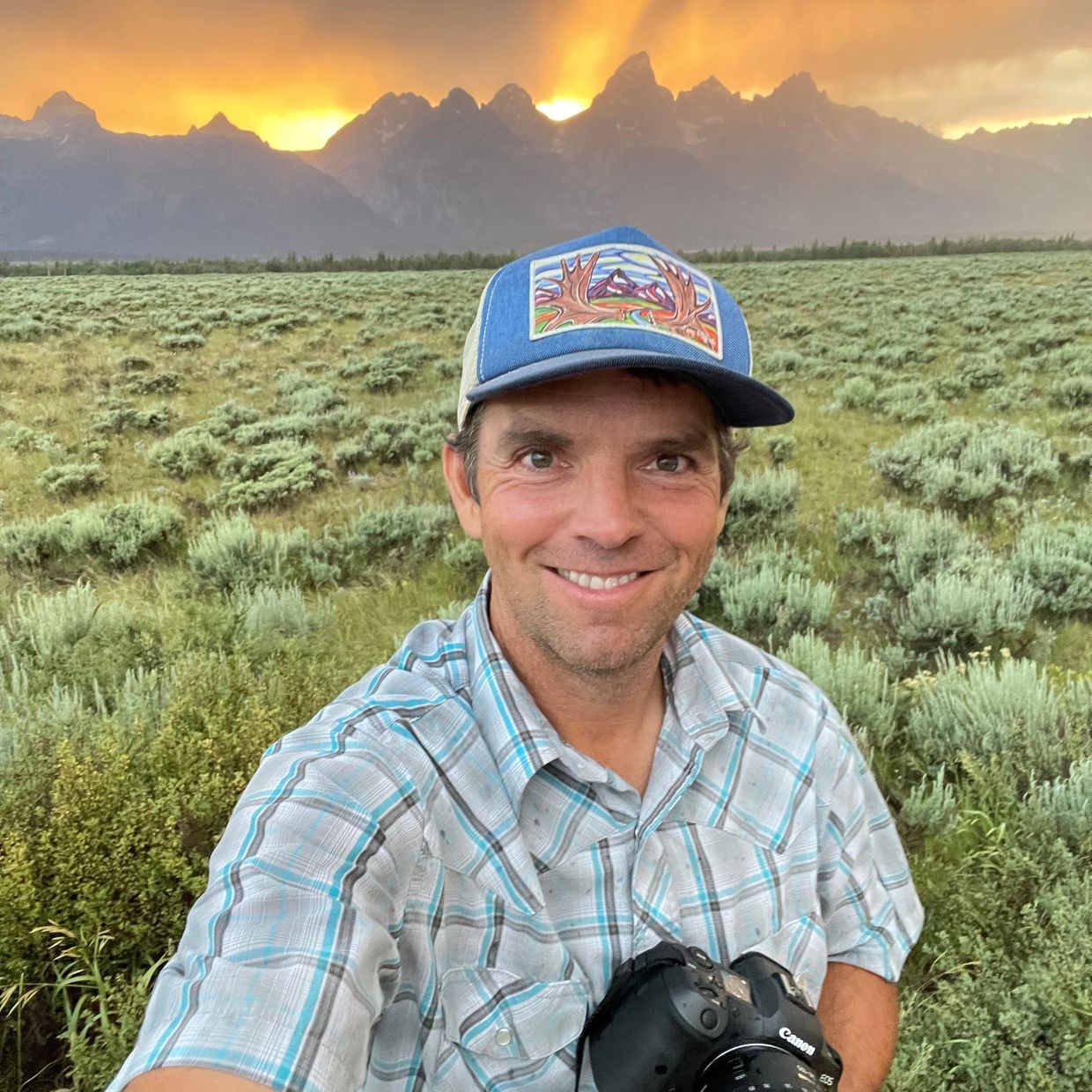 Man with camera takes selfie in scenic field at sunset with mountains in background.