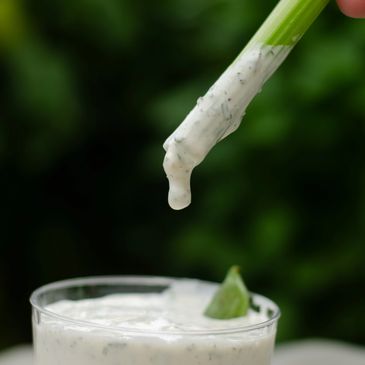 Celery stick dipped in creamy ranch dressing with a glass of dressing below.