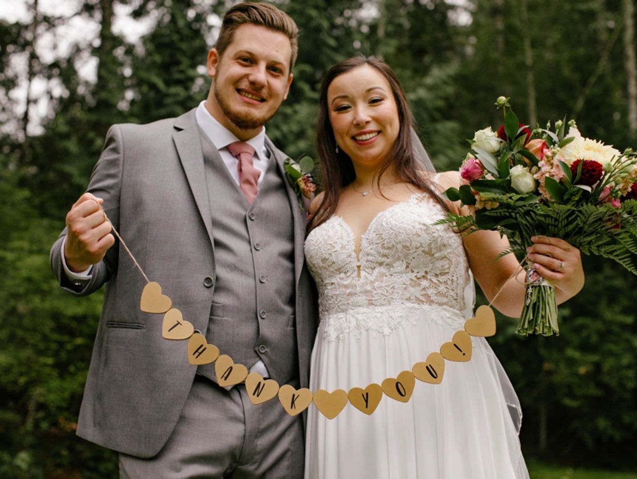 Groom in gray with a pink tie and bride  holding a garland of cut-out hearts that say "thank you!" 