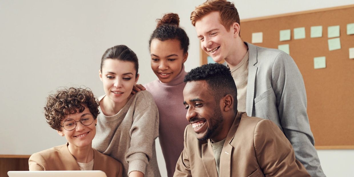 Five people standing in a group, smiling, looking at a laptop screen
