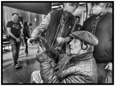 Elderly man with headphones and gloves using a smartphone, surrounded by masked people indoors.