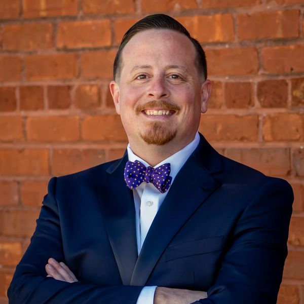 Man in suit with bow tie smiling in front of brick wall.