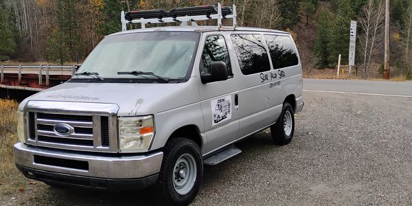 Silver Ford van parked on a gravel road near a wooded area.