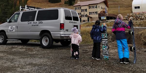 Three children with snowboards near a shuttle van in a mountainous area.