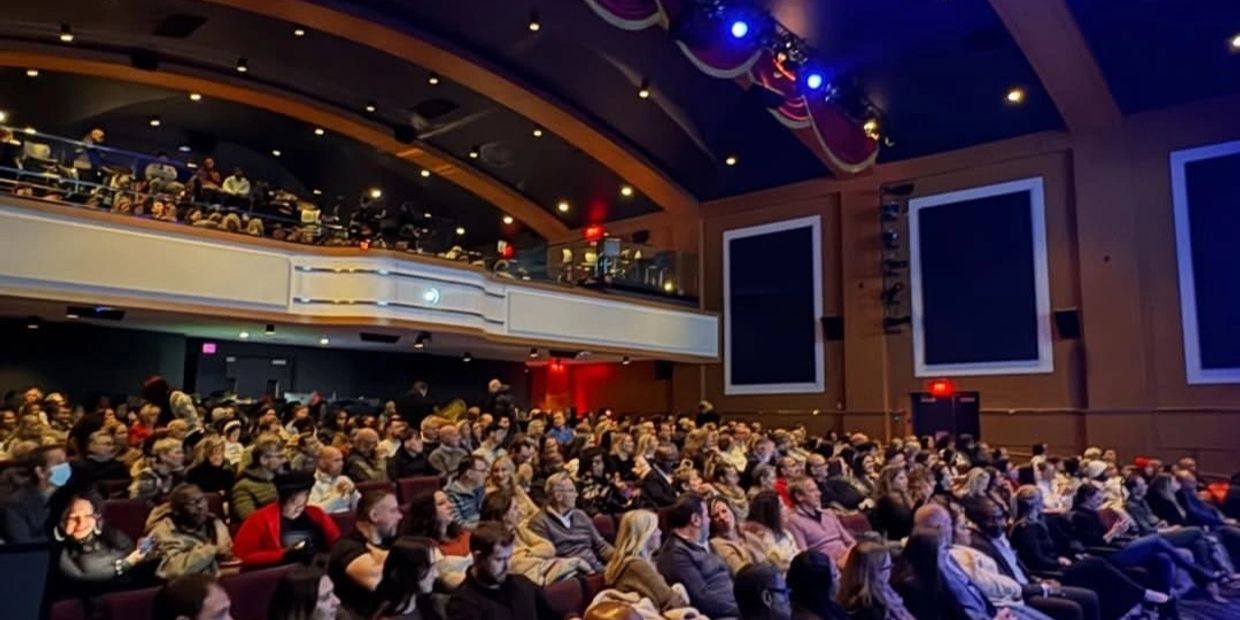 A large audience seated in a theater with a balcony, waiting for a performance to start.