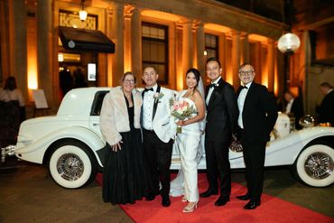 Elegant wedding party posing on red carpet with vintage white car at night.