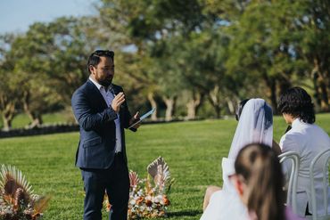 Man in suit speaking at an outdoor wedding ceremony.