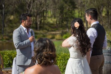 Man in gray suit speaking at a wedding ceremony outdoors.