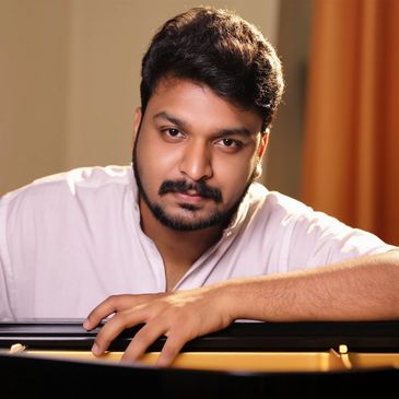 A man in a white shirt leans over a piano, looking thoughtfully at the camera.