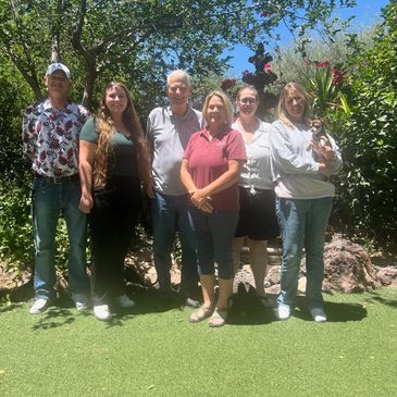 A group of six adults standing outdoors on grass with trees in the background.