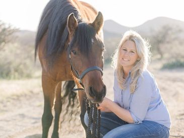 Woman crouching next to a brown horse in a sunny outdoor setting.