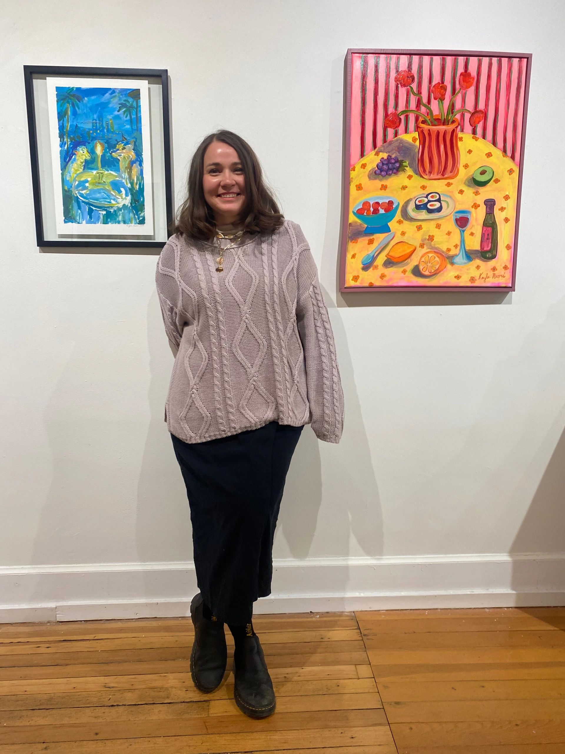 A woman smiling in front of colorful paintings in an art gallery.