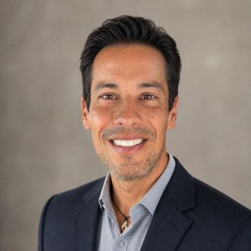 Smiling man in a dark blazer and light shirt against a plain background.