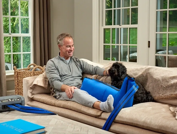 Man relaxing on a couch with a dog wearing a blue recovery suit.