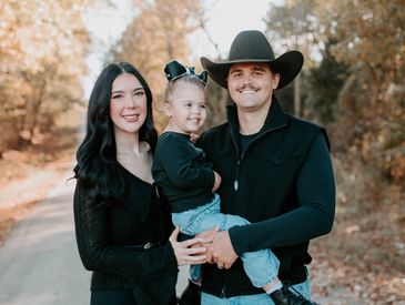 A family of three smiling outdoors in autumn attire.