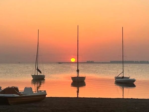 Three sailboats on calm water at sunset with an orange sky.