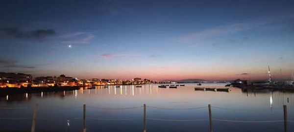 Calm waterfront at dusk with boats and city lights reflecting on the water.