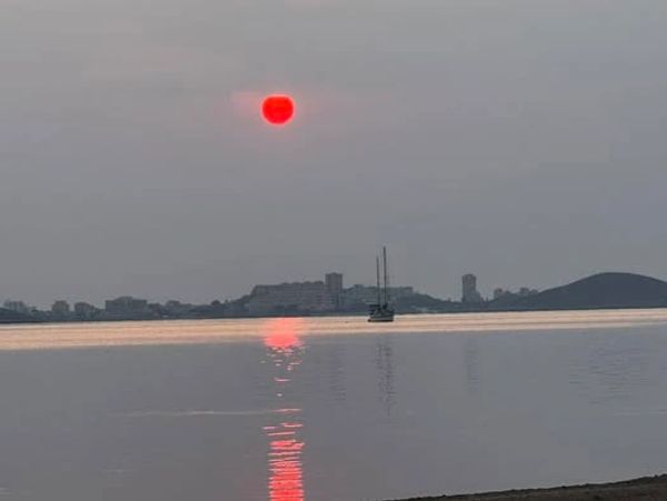 Red sun setting over calm water with a sailboat and silhouetted cityscape.