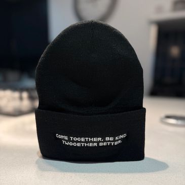 Black beanie with white embroidered text on a light surface.