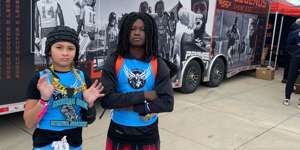 Two young athletes in blue jerseys stand confidently in front of a sports trailer.
