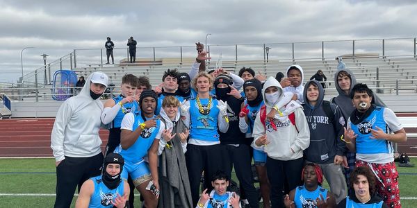 A group of young athletes posing on a football field, some wearing blue jerseys and others in casual clothes.